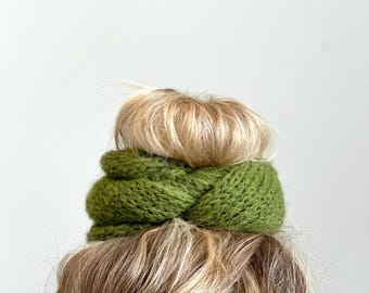 May include: A green knitted hair scrunchie with a twisted design. The scrunchie is worn around a bun of blonde hair.