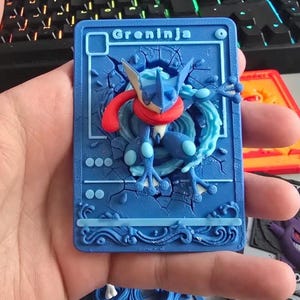 Puede incluir: Una placa rectangular azul impresa en 3D con un relieve del personaje Greninja en el centro. El personaje es azul con una bufanda roja. El fondo de la placa tiene una textura de piedra agrietada y el texto Greninja en la parte superior.