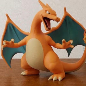Op de afbeelding: Een Charizard-figuur met een oranje lichaam, een crèmekleurige buik, blauwgroene vleugels en een vlam aan het uiteinde van de staart. Het wezen staat in een staande houding met de bek open, zichtbare tanden en klauwen aan de handen en voeten.