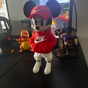 Op de afbeelding: Een gestileerd Mickey Mouse-figuurtje met een rode Nike-honkbalpet, een rode Nike-hoodie, een witte cargobroek en bijpassende witte sneakers. Het personage heeft een strenge gezichtsuitdrukking en grote zwarte oren.