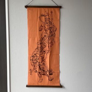 May include: Orange wall hanging featuring a dark brown illustration of two figures. The figures are detailed with intricate lines against a solid orange background. Wooden rods are at the top and bottom for hanging.