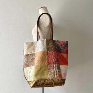 以下が含まれることがあります： 茶色の革の持ち手が付いたパッチワークのトートバッグ。ベージュ、茶色、赤、オレンジの色合いの模様の生地の正方形が特徴です。バッグはマネキンに展示されています。