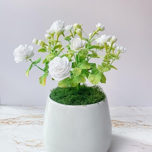 May include: A white ceramic pot with a crocheted flower arrangement. The arrangement features white flowers and green leaves. The pot is filled with green moss. The pot has a smooth, glossy finish and a rounded shape. The flowers are in full bloom.