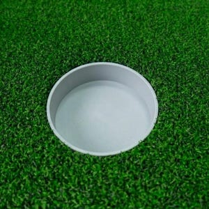 Puede incluir: Un green de golf con un hoyo de golf blanco. La copa está situada en el centro del green, rodeada de césped artificial verde y vibrante. La copa tiene un diseño circular simple.