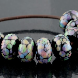Könnte beinhalten: Eine Reihe von sieben runden, schwarzen Glasperlen, die auf einer dünnen braunen Schnur aufgereiht sind. Jede Perle hat ein gesprenkeltes Muster aus pastellfarbenen Punkten in Hellblau, Rosa, Gelb und Grün. Die Zahl 1004 ist hellblau aufgedruckt.