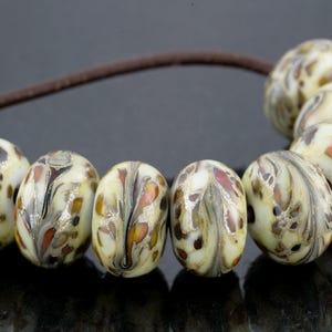 May include: Seven round glass beads with a cream-colored base and swirling patterns of brown, black, and orange, strung on a thin brown cord. The number 1016 is printed in light blue in the upper left corner of the image.