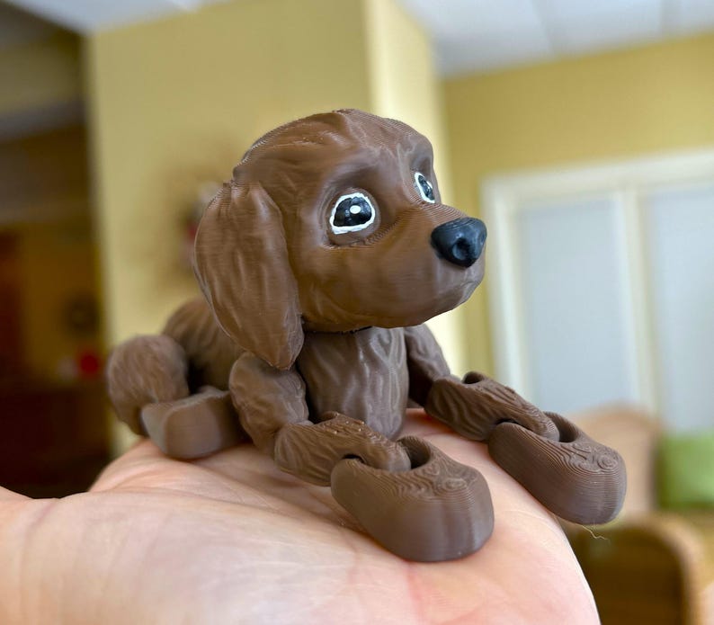 Peut inclure: Un chien en jouet imprim&eacute; en 3D de couleur marron, articul&eacute;, avec de grands yeux et une expression espi&egrave;gle. Le chien est en position assise, les pattes avant tendues.