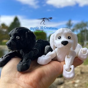 Peut inclure: Deux figurines de chien imprim&eacute;es en 3D, l'une noire et l'autre blanche, sont pos&eacute;es sur une main. Les figurines sont articul&eacute;es et ont une expression espi&egrave;gle. Le texte "Mystic 3D Creatures" est visible en arri&egrave;re-plan.