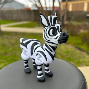 May include: A 3D printed zebra figurine with black and white stripes. The zebra has large eyes and a playful expression. The figurine is standing on a gray surface.