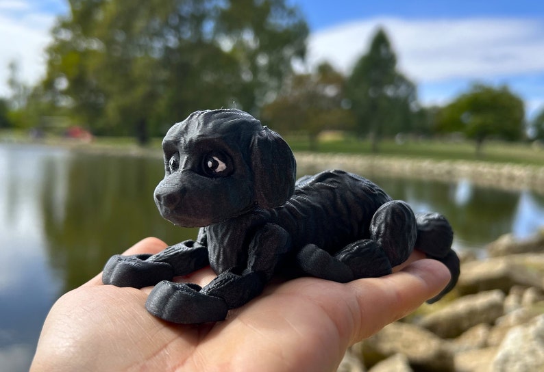 Peut inclure: Une figurine d'un chien en 3D imprim&eacute;e en noir, couch&eacute;e avec la t&ecirc;te tourn&eacute;e sur le c&ocirc;t&eacute;. La figurine a une surface textur&eacute;e et des d&eacute;tails r&eacute;alistes.