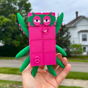 May include: A vibrant pink and green toy figure with a rectangular body, green arms, and a green mask. The figure has a smiling face with two eyes and the number 8 on its side. The toy is held in a person's hand.