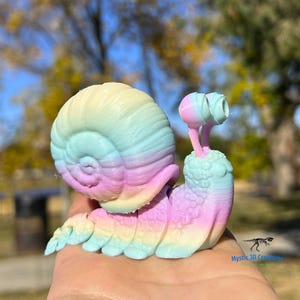 May include: A rainbow-colored 3D printed snail figurine with a detailed shell and textured body. The snail is posed with its head and eyes extended.