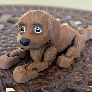 Peut inclure: Un chien en jouet articul&eacute; brun avec des yeux blancs et un nez noir. Le chien est couch&eacute; sur une surface en osier brun.