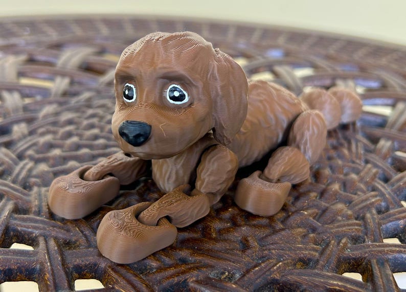 Peut inclure: Un chien en jouet articul&eacute; brun avec des yeux blancs et un nez noir. Le chien est couch&eacute; sur une surface en osier brun.