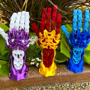 May include: Three articulated hand sculptures in vibrant colors: purple and white, red and gold, and blue. The hands are posed in a gesture, with intricate details and a skeletal design. They are displayed outdoors among greenery.