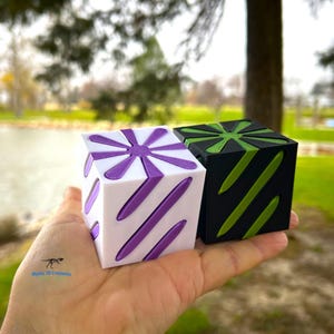 May include: Two small, cube-shaped, 3D printed objects. One is white with purple accents and the other is black with green accents. Both have a starburst design on one side and diagonal stripes on the other.