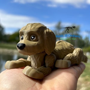 Peut inclure: Une figurine de chien en 3D imprim&eacute;e en brun avec de grands yeux, couch&eacute;e sur une main. La figurine est d&eacute;taill&eacute;e avec de la fourrure et a une apparence r&eacute;aliste. L'arri&egrave;re-plan montre une image floue d'un &eacute;tang et d'arbres.