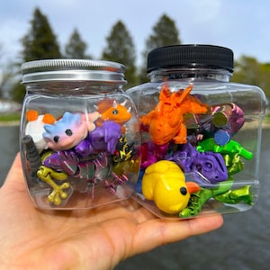 May include: Two clear plastic jars filled with colorful, miniature dragon figurines. One jar has a silver lid, the other a black lid. The dragons are various colors, including purple, orange, yellow, and green. The jars are held in a hand.