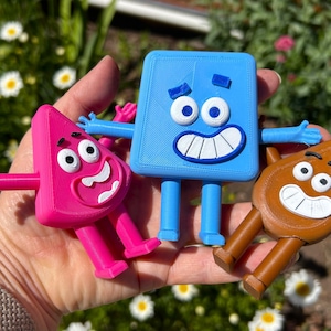 May include: Three brightly coloured, cartoon-style figures with arms and legs. One is pink and triangular, another is blue and square, and the third is brown and teardrop-shaped. Each has a smiling face with white teeth and large eyes. The figures are held in a hand.