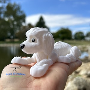 Peut inclure: Figurines de chien blanc imprim&eacute; en 3D allong&eacute;. Le chien a un pelage duveteux et des yeux noirs. La figurine est sur la main d'une personne. Le texte "Mystic 3D Creatures" est visible dans le coin inf&eacute;rieur gauche de l'image.