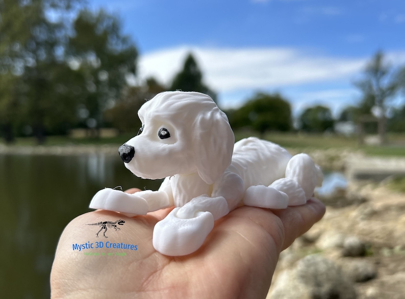 Peut inclure: Figurines de chien blanc imprim&eacute; en 3D allong&eacute;. Le chien a un pelage duveteux et des yeux noirs. La figurine est sur la main d'une personne. Le texte "Mystic 3D Creatures" est visible dans le coin inf&eacute;rieur gauche de l'image.