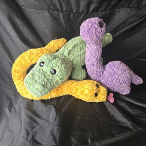May include: Three plush toys made of soft, textured yarn. One is a long, yellow snake with black eyes and a small pink tongue. Another is a green, four-legged creature with black eyes. The third is a purple, long-necked dinosaur-like plush toy.