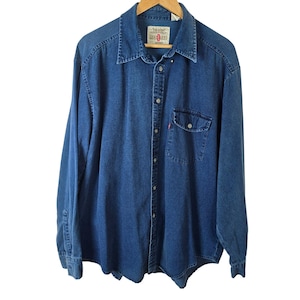 Puede incluir: Camisa vaquera de manga larga en un lavado azul medio, con cuello puntiagudo y un bolsillo en el pecho. La camisa tiene una pequeña etiqueta roja y está colgada de una percha de madera. La camisa está abotonada.