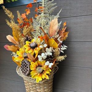 May include: A woven basket filled with an arrangement of artificial sunflowers, orange maple leaves, golden fern fronds, white floral sprigs, pinecones, and a dried lotus seed pod. The decorative display features a warm autumn colour palette.