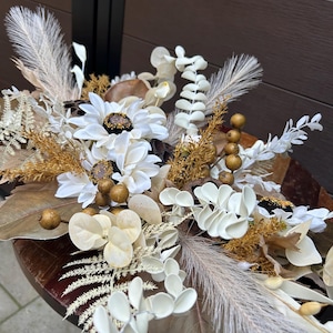 Peut inclure: Un arrangement floral décoratif composé de fleurs blanches semblables à des marguerites aux centres sombres, d'herbe de la pampa crème, de feuilles d'eucalyptus séchées, de brins brun doré et de petites baies rondes brunes.