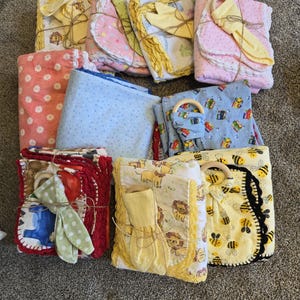 May include: A collection of baby blankets and accessories, including blankets with floral, polka dot, and animal prints. Some sets include wooden teething rings and matching bibs. The blankets are in various colors, including pink, yellow, blue, and red.