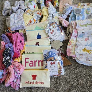 May include: A collection of baby items, including clothing, bibs, hats, and toys. Several small canvas pouches with text and graphics are also visible. A few items are packaged with tags that read "Misc. Felt Pieces."