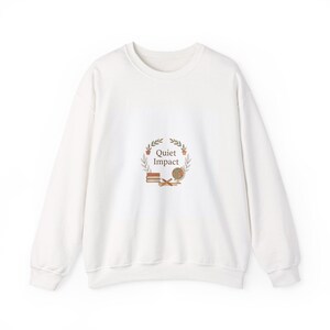 May include: A white sweatshirt with a crew neck. The front features a circular design with the text "Quiet Impact" surrounded by leaves, books, a globe, and small potted plants. The design is in shades of brown and green.