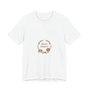 May include: White t-shirt featuring a circular design with the text "Quiet Impact". The design includes a stack of books, a globe, and a pencil, surrounded by a wreath of leaves. The shirt is a classic crew neck style.