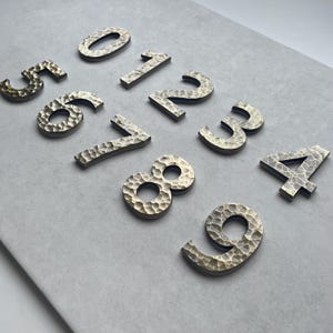 以下が含まれることがあります： ハンマーで叩いたような質感の金色の金属製の家の番号が10個セットになっています。番号は灰色の表面に半円形に配置されています。