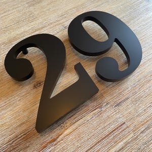 Könnte beinhalten: Schwarze Hausnummern aus Metall, die Ziffern 2 und 9, werden auf einer hellen Holzoberfläche angezeigt. Die Zahlen haben eine moderne, serifenlose Schriftart und sind für den Außenbereich konzipiert. Die Zahlen sind etwa 15 cm hoch.