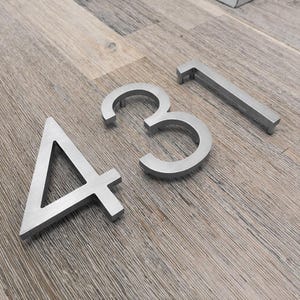 Puede incluir: Números de casa de metal plateado, incluyendo los números 4, 3 y 1, se muestran sobre una superficie de madera. Los números tienen un diseño moderno y un acabado de metal cepillado. Estos números son probablemente para una dirección de casa o edificio.