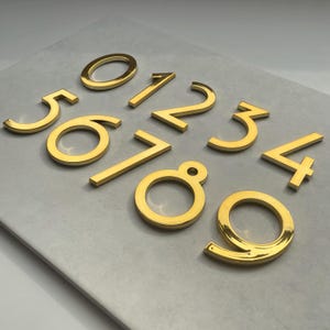 Op de afbeelding: Een set gouden metalen huisnummers, van 0 tot en met 9, op een wit oppervlak.