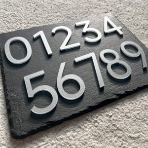 Może przedstawiać: Czarna tabliczka ze łupka z srebrnymi metalowymi cyframi 0123456789. Cyfry są wypukłe i mają nowoczesny, minimalistyczny design.