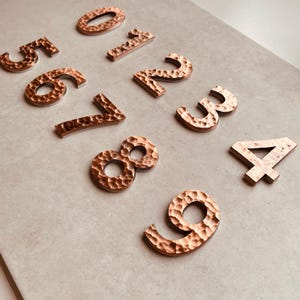May include: Set of ten copper house numbers, each with a hammered texture. The numbers are arranged in a semi-circle on a light grey surface.