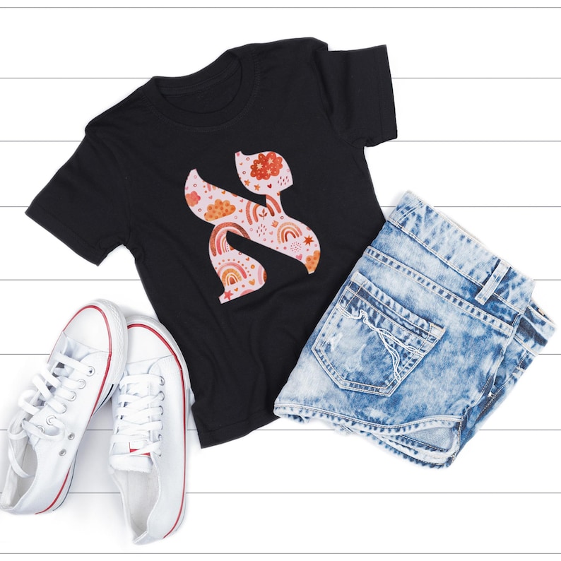May include: A black t-shirt featuring a colorful, floral-patterned letter "S" design with rainbows and clouds. The shirt is paired with acid-wash denim shorts and white canvas sneakers with red accents, all arranged on a white wooden surface.