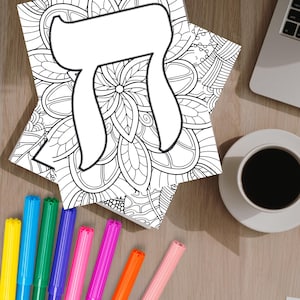 May include: A black and white coloring book page featuring a large Hebrew letter 'Chai' surrounded by intricate floral designs. The page is lying on a wooden table with a cup of coffee and a set of colorful markers.