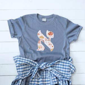 May include: A light blue t-shirt featuring a colorful, whimsical design of the Hebrew letter 'Kaf'. The design incorporates rainbows, clouds, and stars in shades of orange, yellow, and pink. A blue and white plaid shirt is tied at the waist.