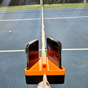 May include: An orange and black phone holder attached to a tennis net post, holding two smartphones. The tennis court is blue with white lines, and a tennis ball is visible in the background.