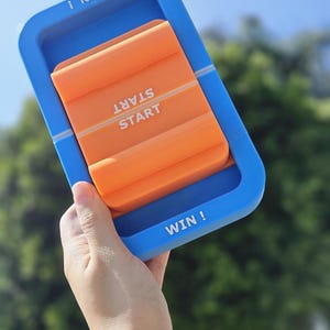 May include: A blue and orange plastic game with the words "WIN!" and "START" on it. The game is being held in a hand.