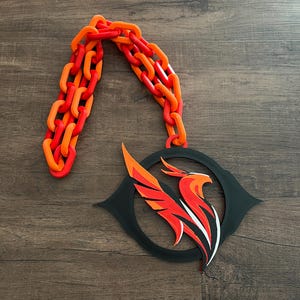 May include: A decorative piece featuring a stylized phoenix design in red, orange, and white, set within a black circular frame. A chain in matching red and orange hues is attached to the top, creating a striking visual contrast.
