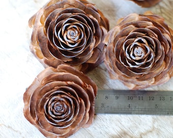 Pode incluir: Quatro rosetas de pinhas marrons, que se assemelham a flores, dispostas sobre uma superfície de madeira. Uma régua é visível no canto inferior direito.