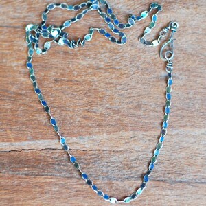 May include: A silver chain necklace with blue and green oval links. The chain is on a wooden surface.