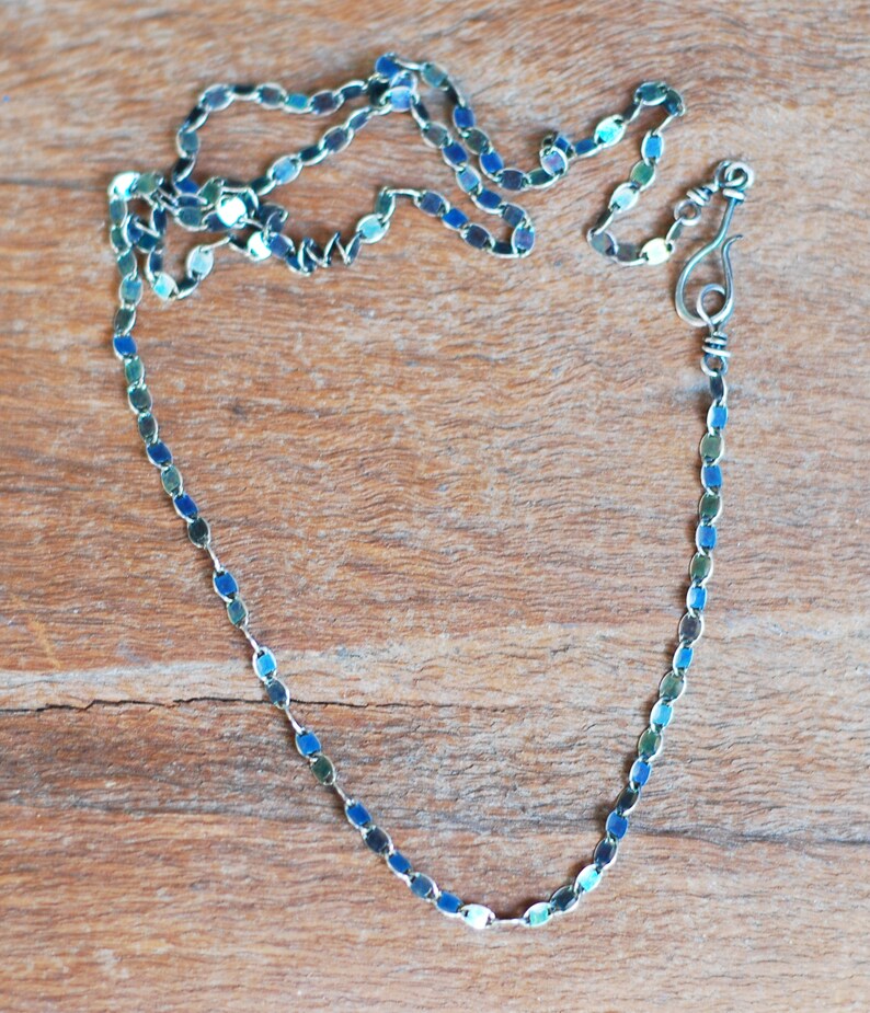 May include: A silver chain necklace with blue and green oval links. The chain is on a wooden surface.