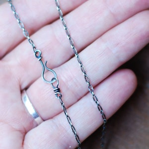 May include: Close-up of a delicate silver necklace with a unique hook clasp. The chain features a repeating pattern of small, textured links. The necklace is displayed against a neutral background, highlighting its intricate design.