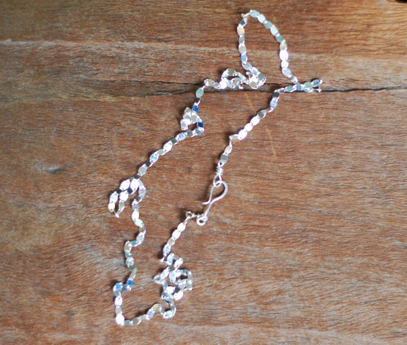 May include: A silver chain necklace with a delicate, flat, oval link design. The chain is on a wooden surface.
